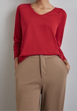 Person wearing a red long-sleeve top tucked into beige high-waist pants with hands in pockets, against a plain light wall.