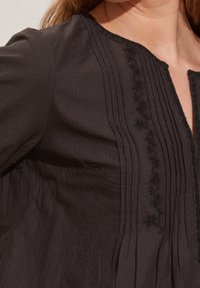 Blusa nera in cotone con pieghe verticali, un design floreale ricamato lungo il collo e una vestibilità morbida con apertura a V.