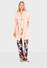 Accappatoio in felpa rosa chiaro con cintura e tre grandi bottoni. Abbinato a pantaloni con motivo floreale, caratterizzati da grandi fiori blu e borgogna su uno sfondo beige.