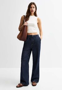Jean en denim foncé à jambes larges associés à un débardeur blanc ajusté à côtes. Un sac à bandoulière marron et des sandales marron complètent le look.
