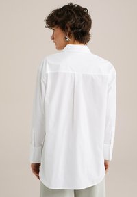 Biela košeľa na gombíky s dlhými rukávmi a voľným strihom. Obsahuje mierne zaoblený lem, zadný rázporok a detaily na manžetách.