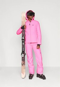 Erksinise roosa veekindlast materjalist suusajope ja püksid. Modell hoiab suuski koos suusapaeltega. Mustad suusasaapad ja suusaprillid täiendavad komplekti.