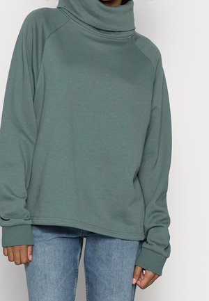Personne portant un sweat-shirt col roulé ample vert discret et un jean bleu clair, debout devant un fond clair uni.