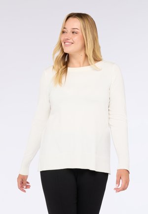 Lächelnde Frau mit blonden Haaren trägt einen weißen Pullover mit langen Ärmeln und schwarze Hosen, steht vor einem schlichten hellen Hintergrund.