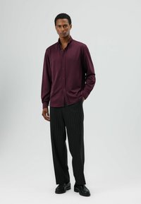 Bordeauxrode blouse met lange mouwen en knopen, met een rechte pasvorm, gecombineerd met zwarte broek met verticale strepen en zwarte veterschoenen.