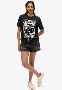 T-shirt grafica nera con stampa bianca che presenta un design di volto, abbinata a pantaloni shorts in denim strappati e sneaker beige.