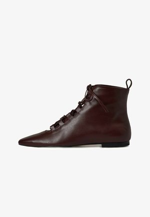 Stivali ankle in pelle marrone con punta affusolata, caratterizzati da un design con lacci e una suola piatta. Texture liscia con una finitura morbida.