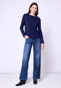 Eine Frau steht mit einer Hand in der Tasche, trägt einen marineblauen Langarmpullover, weite blaue Jeans, schwarze Stiefeletten mit Absatz und silberne Ohrringe.