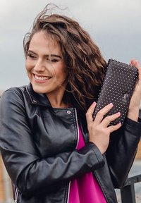 Black wallet with gold polka dots, sleek design, held in hands. Model wears a black leather jacket over a pink shirt, with wavy hair.