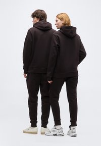 Δύο άτομα στέκονται δίπλα-δίπλα φορώντας σκούρα καφέ φούτερ με κουκούλα και ταιριαστά παντελόνια, στραμμένα με την πλάτη σε απλό λευκό φόντο.