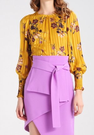 Žena nosící žlutou květovanou blůzu s dlouhým rukávem zastrčenou do vysoké lila zavinovací sukně s páskem na zavázání.