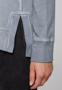 Maglia a maniche lunghe grigio chiaro con polsini e spacco laterale, indossata con pantaloni neri, con una mano rilassata lungo il fianco.