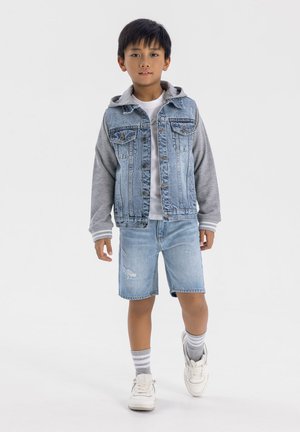 Giacca di jeans con maniche di felpa grigia, t-shirt bianca, pantaloni shorts di jeans strappati e sneakers bianche con calzini a righe grigie.