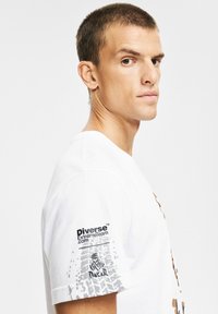 Biała bawełniana koszulka z brązowym nadrukiem graficznym, krótkimi rękawami oraz czarno-szarym logo na lewym ramieniu.