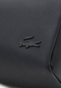 Svart teksturert veske laget av syntetisk materiale med en hevet svart krokodille-logo. Designet er vinkelaktig med en glatt finish.