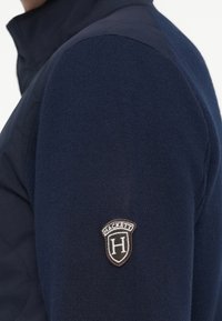 Marineblauwe pullover met een hoge kraag en textuurstof. Voorzien van een logopatch met "HACKETT" en een schildontwerp op de linkermouw.