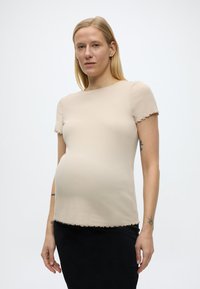 Έγκυος γυναίκα με μακριά ξανθά μαλλιά, φορώντας μπεζ κοντομάνικη μπλούζα και μαύρη φούστα, στέκεται μπροστά σε φωτεινό φόντο.