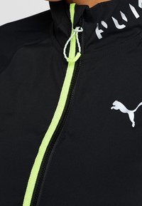 Musta takki neonvihreällä vetoketjun vedolla, valkoinen Puma-logo rinnassa ja osittain näkyvä valkoinen "PUMA"-teksti olkapään lähellä.