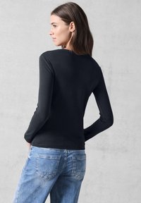 Schwarzes langärmliges Oberteil aus weichem Stoff, mit Rundhalsausschnitt; kombiniert mit hellblauen Jeans, Rückansicht.