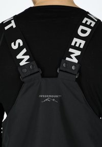 Svart Swedemount bär overall med justerbara axelremmar, med en logotyp och metallknappar på axlarna, tillverkad av slitstarkt tyg.