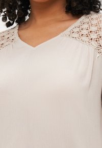 Donna che indossa una blusa beige chiaro con scollatura a V e dettagli in pizzo sulle spalle.