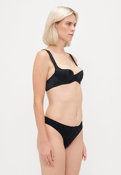 Mulher loira com corte de cabelo à bob, vestindo um soutien preto com aros e uma cueca preta a condizer, de pé de lado contra um fundo branco simples.