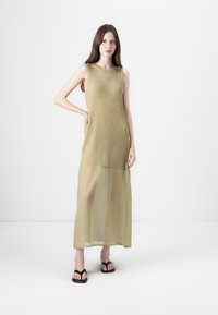 Uten ermer, olivenfarget midi-kjole med teksturert ribbing og et gjennomsiktig nedre panel, parret med svarte flip-flop sandaler.