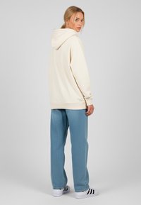 Felpa con cappuccio crema e polsini a costine, abbinata a pantaloni leggeri azzurri con texture e sneakers bianchi con strisce nere.