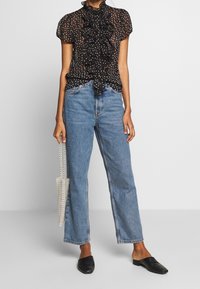 Blusa nera trasparente con pois bianchi, davanti arricciato, maniche corte; abbinata a jeans blu a vita alta e scarpe nere slip-on.