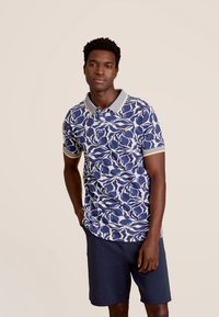 Donkerblauw poloshirt met een wit en blauw abstract patroon, voorzien van een grijze kraag en gele accenten op de manchetten. Draagt bij navy shorts.