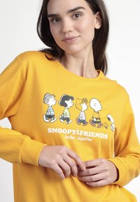 Mujer con sudadera amarilla con Snoopy y los personajes de Peanuts caminando, con el texto "SNOOPY & FRIENDS mejor juntos" en la parte frontal.