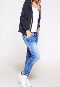 Svart lätt bomberjacka med gul dragkedja, vit T-shirt, ljusblå jeans och vita sneakers med blå detaljer.