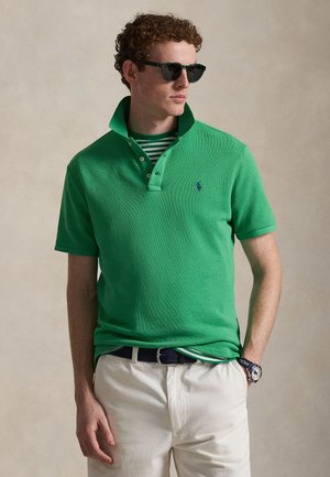 Bărbat purtând tricou polo verde cu guler ridicat peste o cămașă în dungi, pantaloni bej, curea neagră, ochelari de soare și ceas de mână, stând cu o mână în buzunar.