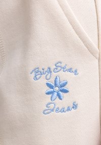 Sinine tikitud lill koos "Big Star Jeans" tekstiga beeži kootud kangal, rõhutades brändi logo detaili rõivastel.