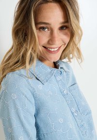 Chemise en denim bleu clair avec broderies florales, col pointu et poche avant. Présente un tissu texturé et des motifs floraux détaillés.