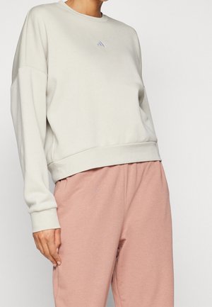 Persoon draagt een lichtbeige sweatshirt met een klein logo op de borst en losse roze broek tegen een effen achtergrond.