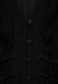 Top de tela negra con escote en V, seis botones negros en la parte delantera y textura fruncida a lo largo de los lados. Material ligero y texturizado.