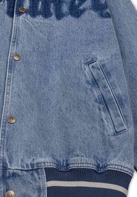 Chaqueta bomber de denim en azul claro, con cierre frontal de botones a presión, bolsillos laterales y puños acanalados en color navy con rayas blancas. Acabados desgastados visibles.