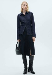 Chemise en denim foncé avec boutons à l'avant, associée à une jupe en denim à mi-cuisse avec une fente avant. Bottes noires jusqu'aux genoux et un sac noir.