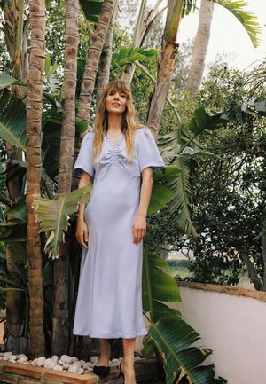 Naine hele sinise midi kleidi ja mustade kontsadega seisab kõrgete palmipuude ja suurte roheliste lehtede vahel õues.