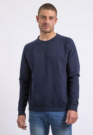 Homme portant un sweat-shirt bleu marine avec un texte brodé discret, associé à un jean bleu clair, debout devant un fond blanc uni.