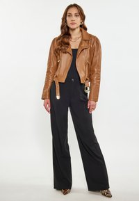 Giacca da biker in pelle marrone con chiusure a zip e dettagli sulle spalle, abbinata a pantaloni neri a gamba larga e scarpe a fantasia.