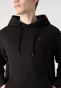 Melna kokvilnas kapuce ar priekšējo kenguru kabatu, regulējamiem aukliņām un mazu sarkano "ck" logo uz krūšu daļas.