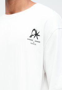 Weißes Langarm-T-Shirt mit schwarzem Grafiklogo und Text auf der oberen linken Brust, aus glattem Baumwollmaterial gefertigt.