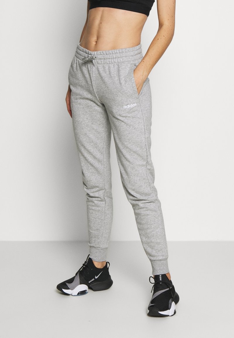 Femme portant un pantalon de survêtement Adidas gris clair, un soutien-gorge de sport noir et des baskets de sport noires, debout avec une main dans la poche.