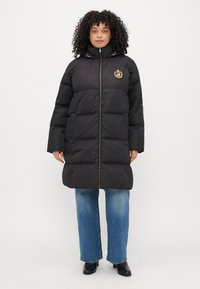 Čierna puffer bunda s vysokým golierom, s logom na nášivke, predným zipsom a veľkými prešívanými časťami. Spárovaná so širokými modrými džínsami.