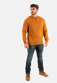 Sudadera naranja con puños y dobladillo acanalados, llevada sobre vaqueros azules y botas marrones. La tela parece suave y tiene una forma ajustada.
