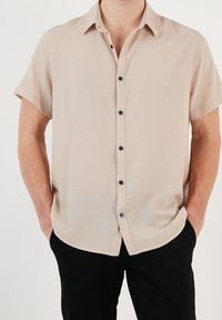 Camisa de manga corta color beige con tela texturizada y cierre de botones al frente, con botones azul oscuro y dobladillo redondeado.