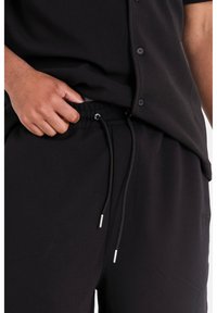 Shorts noirs avec une ceinture à cordon ajustable et des aglets en argent, associés à une chemise à boutons sombre, détail de texture visible.