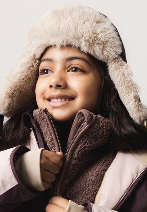 Enfant souriant portant un chapeau d'hiver en fourrure et une veste zippée, tenant la fermeture éclair de la veste avec les deux mains.
